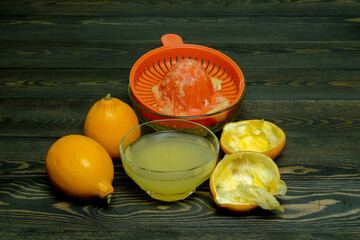 squeezed juice with a hand juicer in a glass cup, two whole lemons and two squeezed lemon peels on a dark, coarse wooden background from the boards
