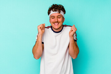 Young sport caucasian man isolated on blue background raising fist, feeling happy and successful. Victory concept.