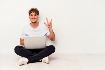 Young caucasian man sitting on the floor holding on laptop isolated on white background showing a horns gesture as a revolution concept.