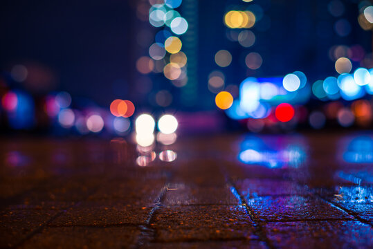 Rainy Night In The City. The Headlights Of The Car Going Through The Parking Lot. Light From The Windows Of The House Opposite. Close Up View From The Pavement Level.