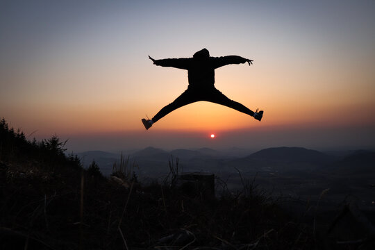 Silhouette of a teenager and athlete jumping on a tree stump and doing a straddle vault, splits at sunset in the European mountains. Training in natural conditions. Self weight training. Coordination