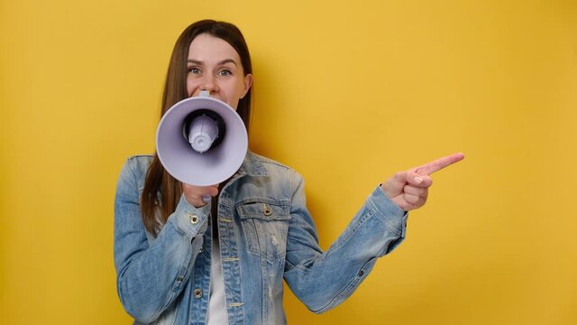 Excited shocked young woman 20s pointing index finger aside screaming in megaphone, dressed in denim jacket, posing isolated on yellow studio background. People lifestyle concept. Mock up copy space