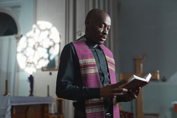 African young priest reading the Bible during mass while standing near the altar in church
