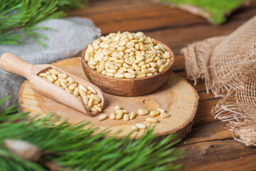 A plate and a scoop filled with pine nuts are on a wooden board on the table. Nearby are pine branches, gray fabric. 