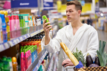 Sceptic male in bathrobe reading information about shampoo, choosing the best one in store