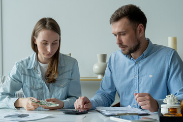 Young married couple sitting at living room table, studying papers, makes calculations on a calculator, studies the family budget