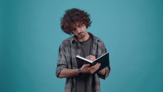 The charismatic, serious young man makes thoughtful notes and looks thoughtfully to one side, what else to write . Isolated on a turquoise background. Concept of life. People's emotions. 4k portrait