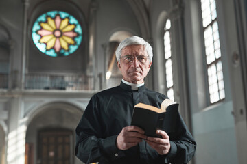 Portrait of senior priest holding the Bible and looking at camera while standing in the church - obrazy, fototapety, plakaty