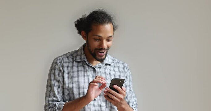 Emotional happy young african ethnic hipster man looking at telephone screen, feeling excited of getting message with amazing news, celebrating online lottery win isolated on white studio background.