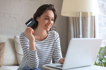 Happy natural brunette using credit card and laptop in the living room.