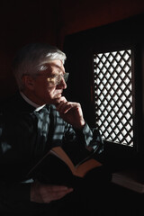 Serious senior priest sitting with Bible in confessional in the church