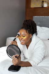 Relax and beauty procedures. Gorgeous African woman lying on bed in cozy room, with golden eye patches under eyes