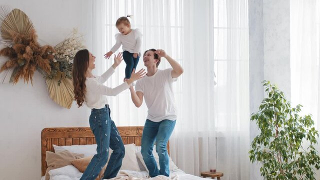 Young family Caucasian parents with small daughter playing at home on bed, strong father man holding baby infant little girl with one hand acrobatic pose mom catches child laughing throws kid into air