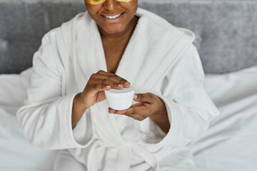 Concept of cosmetology and dermatology. happy black woman sitting on bed at home, using cream