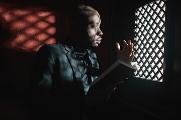 African young priest reading the Bible while sitting in confessional - obrazy, fototapety, plakaty