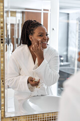 Fototapeta premium Cute afro american lady in white robe enjoy taking care about herself, applying moisturizing cream