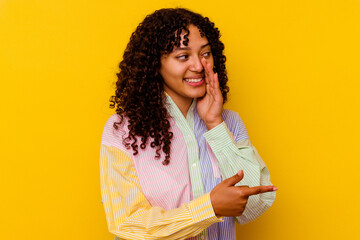 Young mixed race woman isolated on yellow background saying a gossip, pointing to side reporting something.