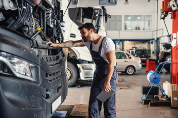Hardworking tattooed bearded mechanic leaning on truck and checking on motor while standing in...