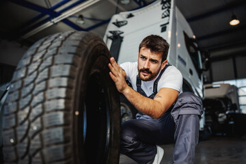 Young dedicated hardworking mechanic crouching in garage of import and export firm and preparing to change tire on truck. - obrazy, fototapety, plakaty