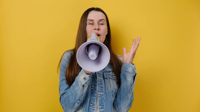 Excited shocked funny young caucasian female screaming in megaphone spreading hands, dressed in denim jacket, posing isolated on yellow color background studio. People emotion lifestyle concept