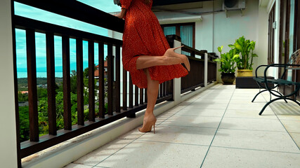 A girl stands in the summer on a balcony in a red dress and heels lifting one leg