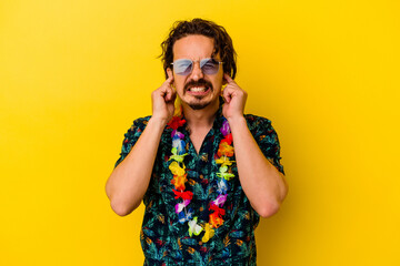 Young caucasian man wearing a hawaiian necklace isolated on yellow background covering ears with hands.