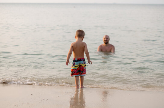 A Blondy-haired Boy Of Three Years Old Stands By The Water And Is Afraid To Go Into The Sea. Dad Calls The Child To Swim In The Ocean