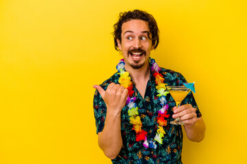 Young caucasian man wearing a hawaiian necklace holding a cocktail isolated on yellow background points with thumb finger away, laughing and carefree.