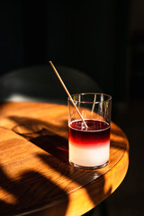 A sweet and sour refreshing blackberry gin bramble cocktail in a highball glass without ice in direct hard sunlight with long shadows, on a wooden table, vertical photo with copy space