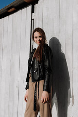 Smiling young woman in black leather jacket standing in front of white wooden wall. Having fun outside. Urban style and street fashion. Girl in casual outfit looking at camera
