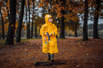 A little boy in a yellow raincoat and rubber boots plays on the street with a flashlight in an autumn park in yellow leaves. Portrait of a little happy and cheerful preschooler