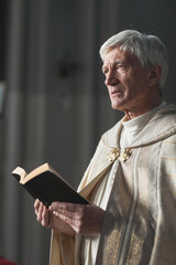 Senior priest standing with Bible and reading prayers during wedding in the church