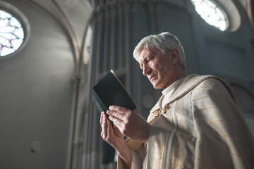 Senior priest in formal costume reading prayers while holding ceremony in the church