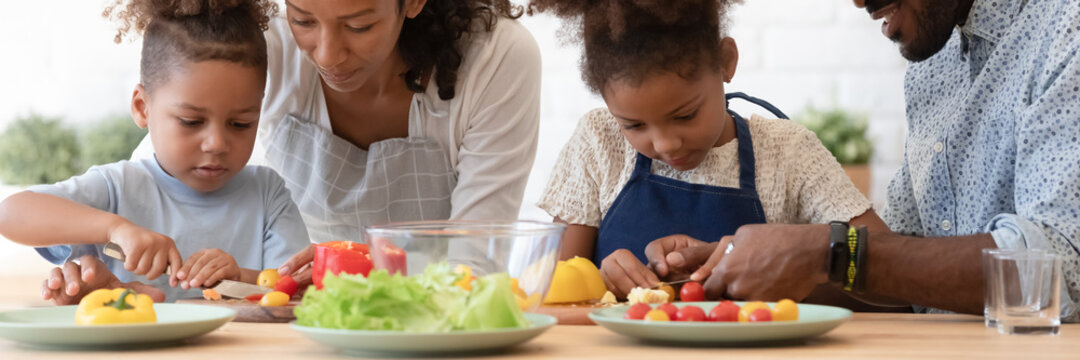 Happy African American parents teaching sibling kids to cook, slicing fresh vegetables, peppers, tomatoes for salad on kitchen table or counter. Family healthy eating, organic food concept. Banner