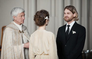 Young couple standing opposite each other and giving prayers at wedding ceremony with priest