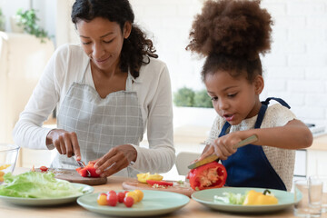 Happy mommy and cute daughter girl in aprons cooking together in kitchen. Mom and kid slicing fresh vegetables for salad, preparing healthy organic meal and talking. Family eating at home concept