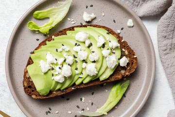 Whole grain rye bread toast with goat cheese and avocado on white stone table background. Healthy avocado open sandwich for breakfast or lunch. Flat lay, top view, close up