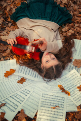 A beautiful girl lies with a violin on sheets with notes among the autumn orange foliage in the forest. She plays the violin