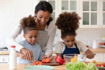 Happy mom teaching two preschooler kids to make vegetarian meal, slicing fresh pepper for salad....