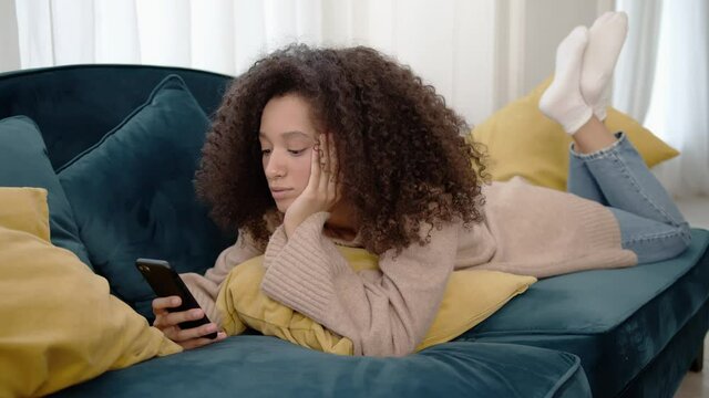 Portrait of african woman using phone, chilling on a long sofa in home living room. Young curly hair woman in beige sweater, smiling and laughing, typing on the mobile phone