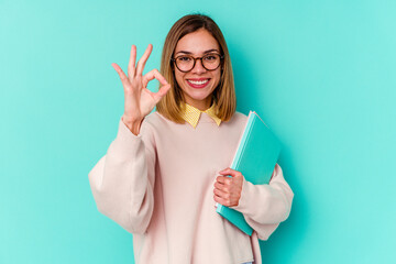 Young student woman holding books isolated on blue background cheerful and confident showing ok gesture.
