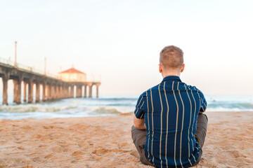 A young blonde man rests on the sand in front of a pier on Manhattan Beach, morning dawn, Los Angeles