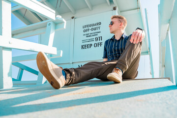 Handsome young blonde man in shirt poses on light blue lifeguard tower on Los Angeles beach