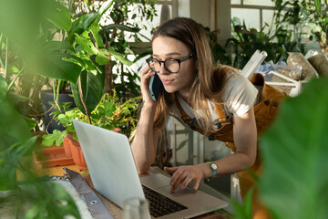 Freelance female work from home, woman gardener use laptop while talking with client on smartphone....