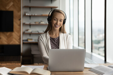 Smiling young Caucasian female student in earphones look at laptop screen study learn online on gadget. Happy millennial woman in headphones work distant talk on video webcam zoom call on computer.