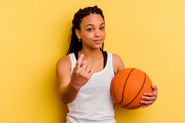 Young african american woman playing basketball isolated on yellow background pointing with finger at you as if inviting come closer.
