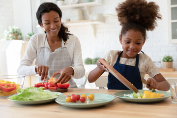 Happy mother and kid sharing cooking chores, preparing salad for dinner together, slicing fresh vegetables on chopping boards, talking and laughing. Family eating at home, healthy food concept