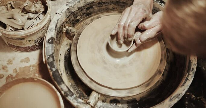 Close up of hands working clay on potter's wheel. Potter shapes the clay product with pottery tools on the potter's wheel, top view, craft factory authentic.