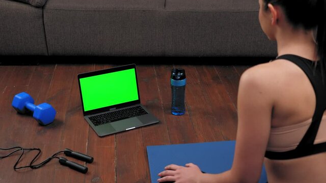 Green screen mock up chroma key monitor laptop concept: Fit woman in sportswear sits on yoga mat looks computer display, tells fitness trainer online video call webcam chat, distance sport course