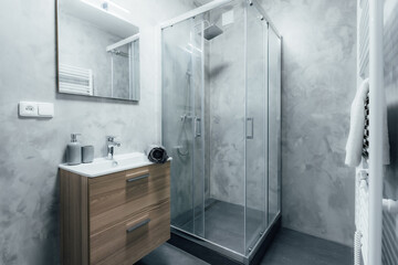 Contemporary bathroom with shower and sink with wooden cabinet under it and mirror with integrated light. All walls are painted with concrete trowel in gray. Whole space gives modern impression. - obrazy, fototapety, plakaty